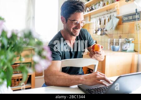 Heureux homme assis à la table dans la cuisine avec ordinateur portable Banque D'Images