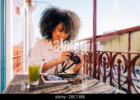 Jeune femme souriante avec des cheveux bouclés assis à la table sur le balcon avec smoothie et appareil photo Banque D'Images