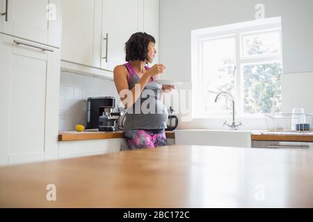 Femme enceinte réfléchie qui mange dans la cuisine avec une fenêtre donnant sur la fenêtre Banque D'Images