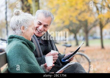 Couple senior souriant lisant le journal et buvant du café sur banc dans le parc d'automne Banque D'Images