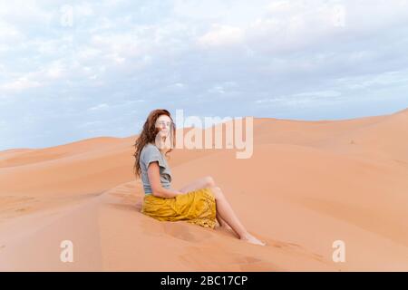 Portrait de jeune femme à tête rouge assise dans une dune de sable dans le désert du Sahara, Merzouga, Maroc Banque D'Images