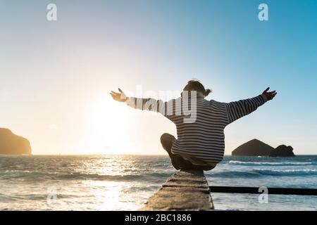 Homme assis sur la côte au coucher du soleil avec des armes étirées, île de Sao Miguel, Açores, Portugal Banque D'Images