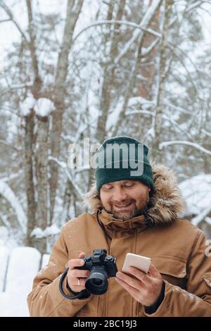 Portrait de l'homme détendu avec appareil photo numérique et téléphone portable en forêt d'hiver Banque D'Images