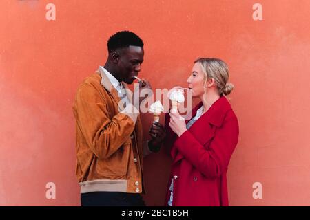 Jeune couple debout sur un mur orange mangeant de la glace Banque D'Images
