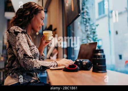 Jeune femme avec un appareil photo utilisant un ordinateur portable dans un café derrière le panneau de fenêtre Banque D'Images