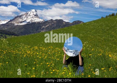 Femme assise dans un pré, se cachant derrière le miroir, reflétant les montagnes de Val Badia, Alto Adige, Italie Banque D'Images