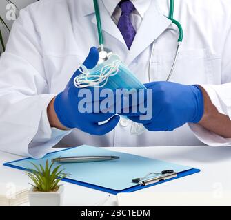 médecin en manteau blanc et gants en latex bleu assis sur un bureau blanc et montre une pile de masques médicaux jetables, agent de protection contre les virus et Banque D'Images