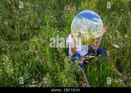 Femme assise dans un pré, se cachant derrière le miroir, reflétant la nature Banque D'Images