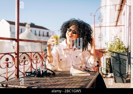 Jeune femme souriante avec des cheveux bouclés assis à la table sur le balcon avec smoothie et appareil photo Banque D'Images