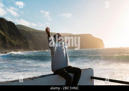 Homme assis sur la côte au coucher du soleil avec des armes étirées, île de Sao Miguel, Açores, Portugal Banque D'Images