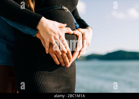 Les parents expectifs affectueux façonnent un cœur avec leurs mains sur le ventre de bébé Banque D'Images
