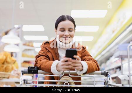 Femme avec smartphone poussant panier dans le supermarché Banque D'Images