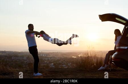 Silhouette d'un père balançant sa fille par les mains, mère assise dans le coffre de voiture en haut de la colline. Belle vue de coucher du soleil sur l'arrière-plan, vue latérale Banque D'Images