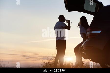 Belle silhouette de couple, homme et femme, ayant romantique temps boire du vin près de la voiture sur la colline au coucher du soleil, ciel du soir sur l'arrière-plan, espace de copie. Femme assise dans un coffre de voiture Banque D'Images