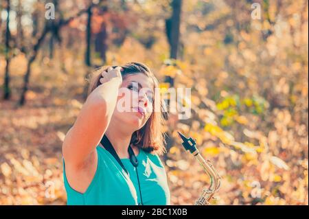 jeune fille brunette très cool avec maquillage dans une robe bleue avec un saxophone dans ses mains redresse ses cheveux, debout dans une forêt jaune d'automne Banque D'Images