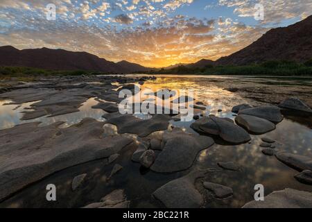 Un beau paysage d'un coucher de soleil doré sur les montagnes et les eaux calmes de la rivière Orange, avec des nuages jaunes spectaculaires reflétant dans l'eau Banque D'Images