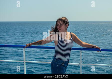 Très belle femme aux cheveux sombres avec un tatouage sur son bras dans un T-shirt rayé et short denim bleu se tient sur la terrasse d'un bateau en mer Banque D'Images