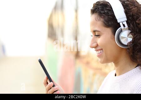 Vue latérale portrait d'une heureuse fille de Latina écoutant de la musique portant des écouteurs et vérifiant le smartphone dans la rue Banque D'Images