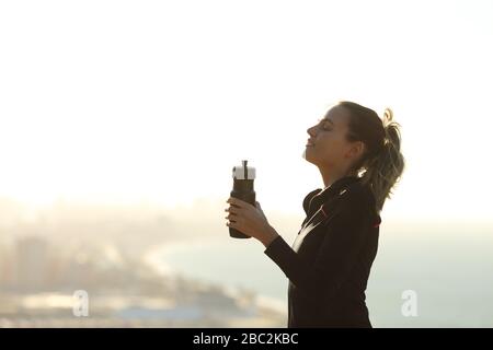 Profil d'une femme de coureur qui se détend respirer de l'air frais tenant une bouteille d'eau avec la ville en arrière-plan