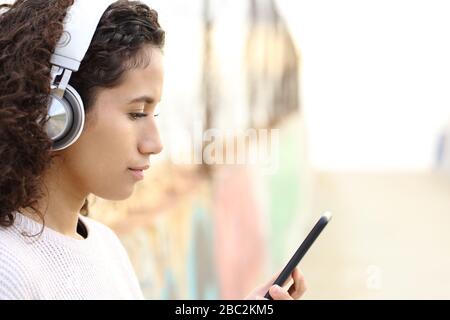 Vue latérale portrait d'une jeune fille latina sérieuse écoutant de la musique portant des écouteurs et regardant le contenu de smartphone dans la rue Banque D'Images