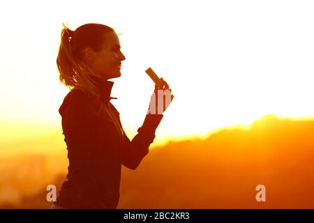 Silhouette d'une femme de coureur qui mangeait un bar énergétique après avoir courir au coucher du soleil dans la montagne Banque D'Images