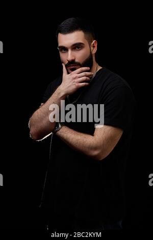 un portrait dramatique d'un jeune homme sérieux, musicien, chanteur, rappeur avec une barbe en vêtements noirs sur un fond noir isolé, il pense attention Banque D'Images
