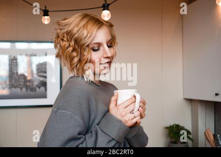 Gros plan portrait d'une belle femme avec une tasse de café. Jolie et heureuse blonde dans le sweat-shirt boit du café. Séjour à la maison - maison confortable Banque D'Images