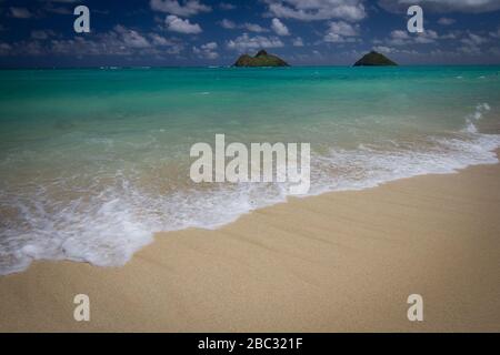 Deux îles au loin, en dessous de nuages blancs puffés, nagent dans les eaux d'aigue-marine au large de la côte d'Oahu, Hawaï. Un doux surf fait des longueurs sur le sable blanc. Banque D'Images