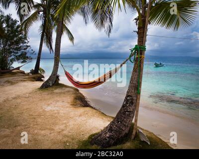 Un hamac se trouve entre deux palmiers à noix de coco au bord de l'eau sur une île de l'océan des Caraïbes. Un bateau de pêche approche à la distance. Banque D'Images