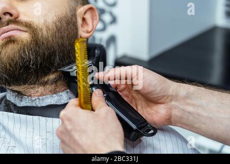 Barbier tailler homme barbu avec machine à raser dans le barbershop. Processus de coiffure. Gros plan d'un stylist coupant la barbe d'un homme barbu. Banque D'Images