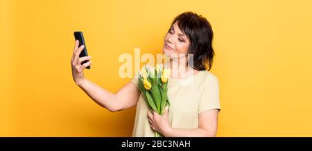 Belle vieille femme avec maquillage professionnel fumé et coiffure tenant des fleurs de tulipe sur le fond rose. Prise d'un appel vidéo par le smartphone Banque D'Images