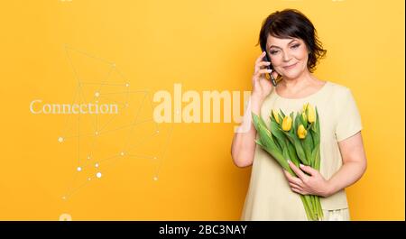 Belle vieille femme avec maquillage professionnel fumé et coiffure tenant des fleurs de tulipe sur le fond rose. Prise d'un appel vidéo par le smartphone Banque D'Images