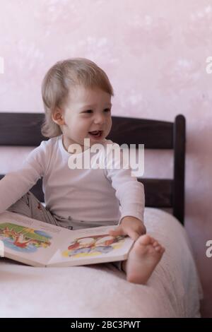 Un petit garçon aux cheveux blonds est assis sur le lit et souriant, tenant un livre dans ses mains. Un petit garçon est assis sur un lit avec un livre dans ses mains pendant Banque D'Images