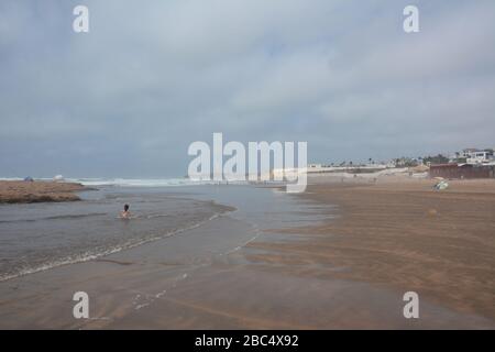 Les habitants de la région se baignent et jouent dans la mer sur la plage publique de Casablanca, au Maroc, avec le phare historique El Hank en arrière-plan. Banque D'Images