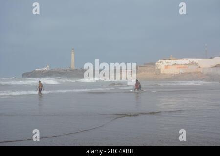 Les habitants de la région se baignent et jouent dans la mer sur la plage publique de Casablanca, au Maroc, avec le phare historique El Hank en arrière-plan. Banque D'Images