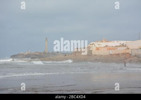 Les habitants de la région se baignent et jouent dans la mer sur la plage publique de Casablanca, au Maroc, avec le phare historique El Hank en arrière-plan. Banque D'Images