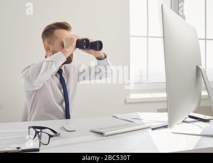 Recherche d'emploi en ligne. Un homme d'affaires dans une chemise blanche regarde à travers des jumelles tout en étant assis à une table avec un ordinateur dans le bureau. Travail de recherche de concept Banque D'Images