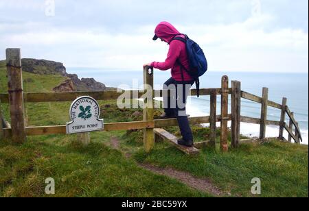 Femme Hiker grimpant sur la tuile en bois par Metal National Trust Signpost pour Steeple point sur le sentier de la côte sud-ouest, North Cornwall, Angleterre, Royaume-Uni. Banque D'Images
