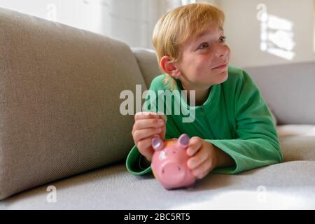 Joyeux jeune beau garçon pensant tout en insérant la pièce de monnaie sur la banque de porc à la maison Banque D'Images