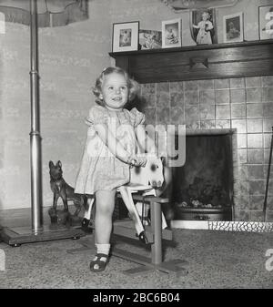 Dans les années 1950, dans un salon, devant un feu de charbon ouvert, une jeune fille heureuse à cheval de son jouet encadré en bois rocking cheval de l'époque, Angleterre, Royaume-Uni. Banque D'Images