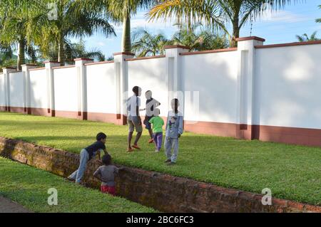 LILONGWE, MALAWI, AFRIQUE - 2 AVRIL 2018: Les enfants locaux se plachent sur le sol avec de l'herbe verte vive près de la clôture et des palmiers blancs, un garçon sourit Banque D'Images