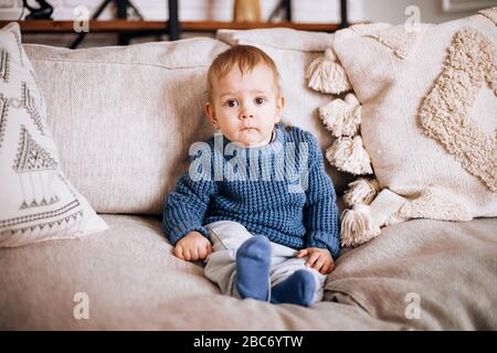 un petit garçon est assis sur un canapé dans un pull bleu et regarde curieusement l'appareil photo Banque D'Images