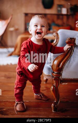 Un petit mignon petit garçon dans une veste et un pantalon rouges, debout à côté d'une chaise douce Banque D'Images