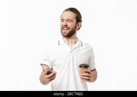 Photo d'un jeune homme émotif barbu posant isolé sur fond de mur blanc à l'aide de la musique d'écoute de téléphone mobile avec écouteurs buvant Banque D'Images