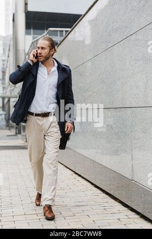 Un jeune homme d'affaires à la fois séduisant et confiant qui marche en plein air dans la rue, porte-documents, parle sur téléphone portable Banque D'Images