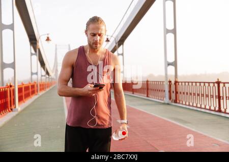 Beau jeune sportif à écouter de la musique avec des écouteurs sans fil sur un pont, tenant le téléphone mobile Banque D'Images