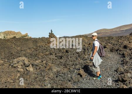 Un jeune homme dans une casquette avec un sac à dos derrière son dos voyage le long d'un itinéraire dans le parc national de Teide. Randonnée pédestre sur le sentier de montagne entouré par ende Banque D'Images