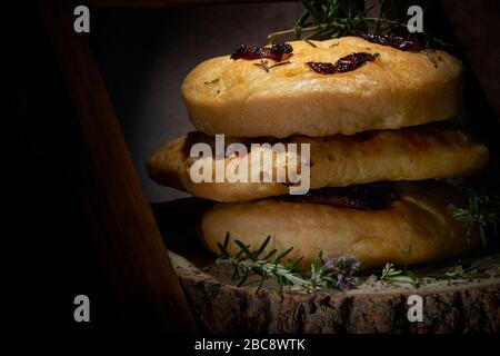 Exposition de pain de Focaccia maison de style italien avec romarin et tomate séchée au soleil. Pain affiché sur bois bark bord studio en basse lumière Banque D'Images