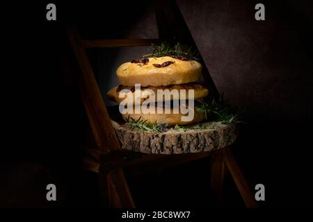 Exposition de pain de Focaccia maison de style italien avec romarin et tomate séchée au soleil. Pain affiché sur bois bark bord studio en basse lumière Banque D'Images