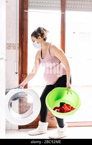 Photo verticale d'une femme enceinte portant un masque dans le visage pour empêcher les virus pendant que fait un lave-linge dans sa maison Banque D'Images
